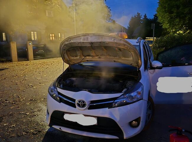 Ein weißer Toyota mit offenem Motorhaube steht rauchend auf der Straße, während es dunkel wird.